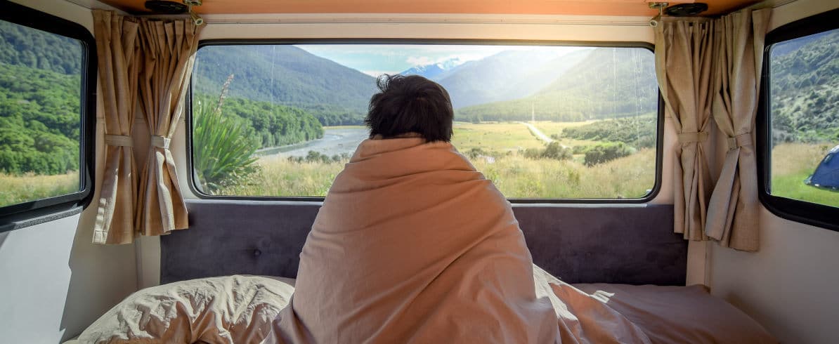 Mann sitzt zugedeckt auf Bett in einem Wohnwagen und schaut aus dem Fenster